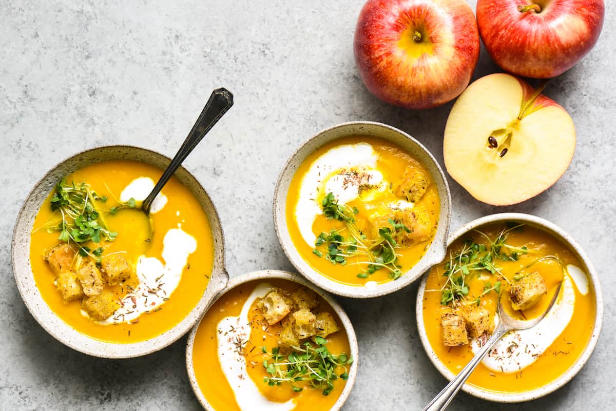 Overhead shot of 4 bowls of carrot and apple soup on gray background, topped with yogurt, herbs and croutons. Apples on table near soup.