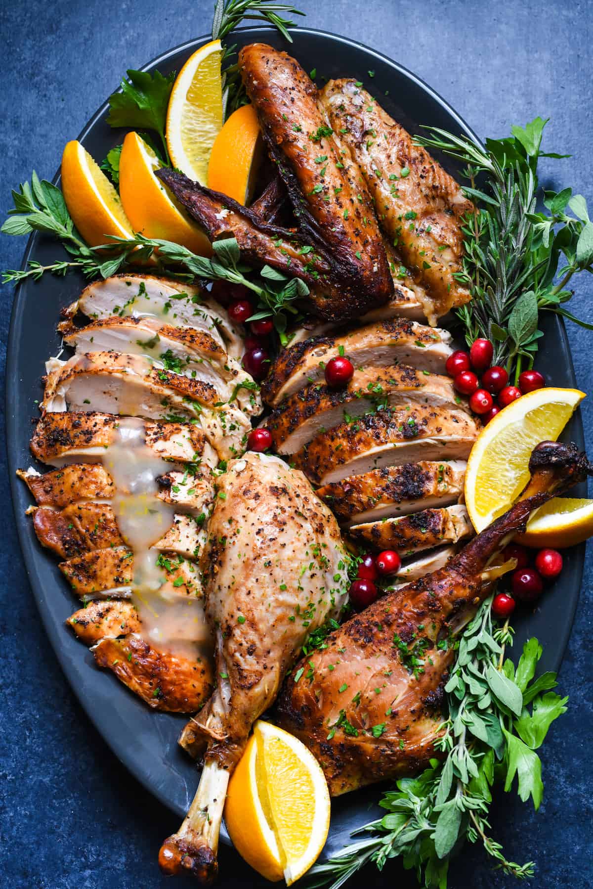 A large dark oval platter with a Cajun turkey that has been roasted and cut up arranged on it, garnished with fresh orange slices, cranberries and herb sprigs.