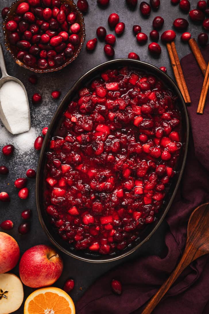 Homemade Cranberry Sauce in a black oval bowl, with the ingredients to make the sauce scattered around the scene.