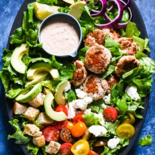 Large platter filled with lettuce, small turkey burgers, red onion, lemon wedges, feta cheese, sliced avocado, sliced grape tomatoes and a small bowl of dressing.