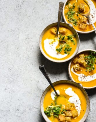 Overhead shot of 4 bowls of carrot and apple soup on gray background, topped with yogurt, herbs and croutons.
