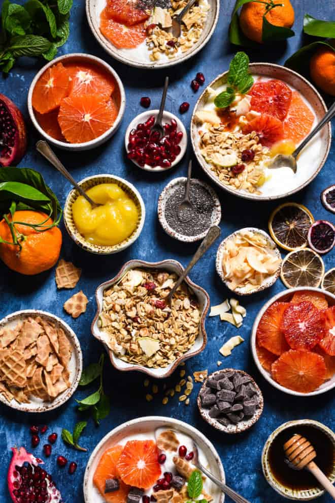 Blue surface covered with a yogurt parfait bar for a brunch buffet, including fruit, seeds, nuts, jams, granola, honey and chocolate.