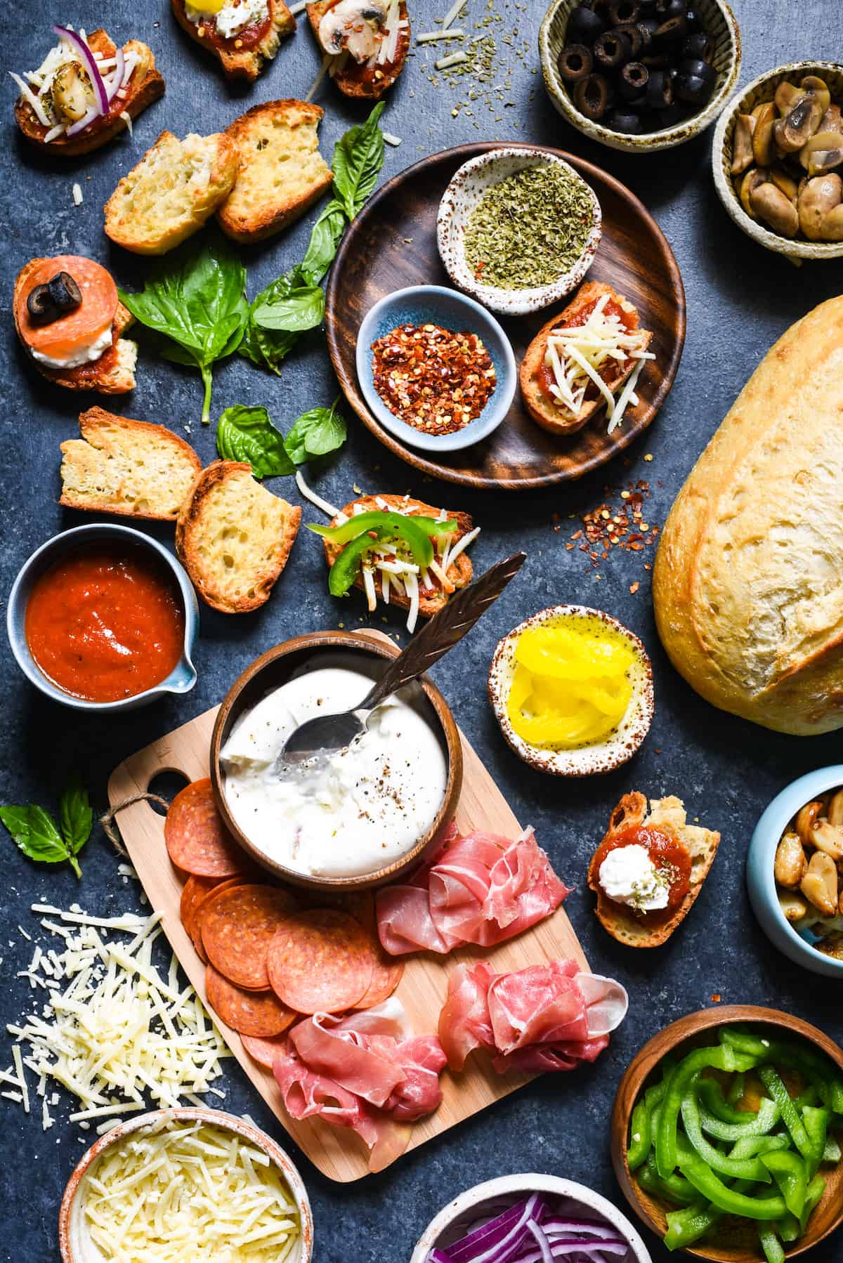 A dark surface filled with the toppings for a crostini pizza bar, including toasted bread, meats, cheese, vegetables and herbs and spices.