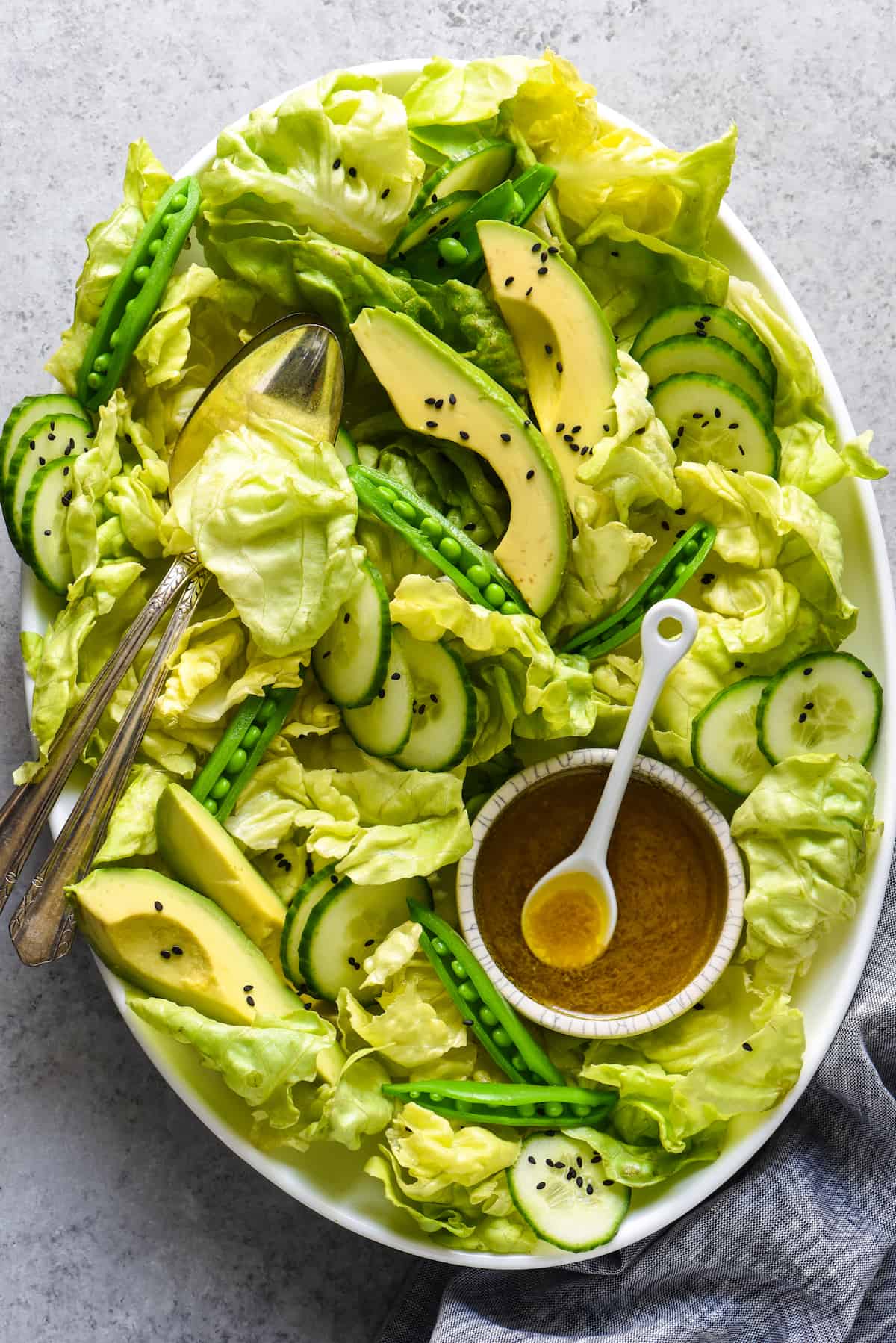 Large platter of green salad with lettuce, avocado, cucumber and peas, and a small bowl of brown salad dressing.