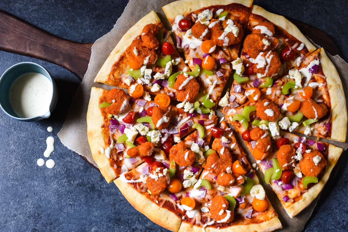 A pizza topped with seafood, spicy sauce, vegetables, blue cheese and ranch dressing on a wooden pizza peel with parchment paper.