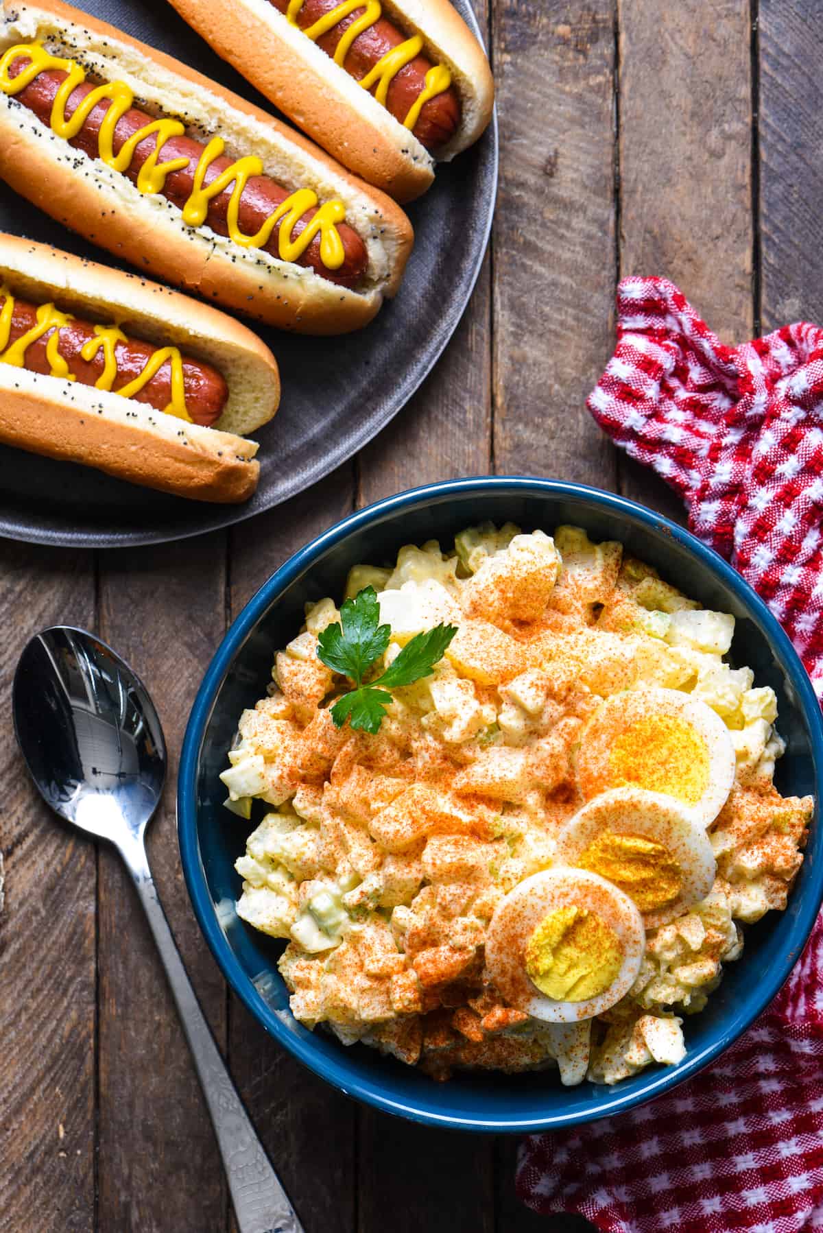 A blue bowl filled with a potato salad recipe with egg, with hot dogs on the side.