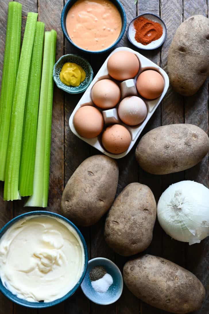 Ingredients on a wooden tabletop, including potatoes, eggs, onion, celery and condiments.