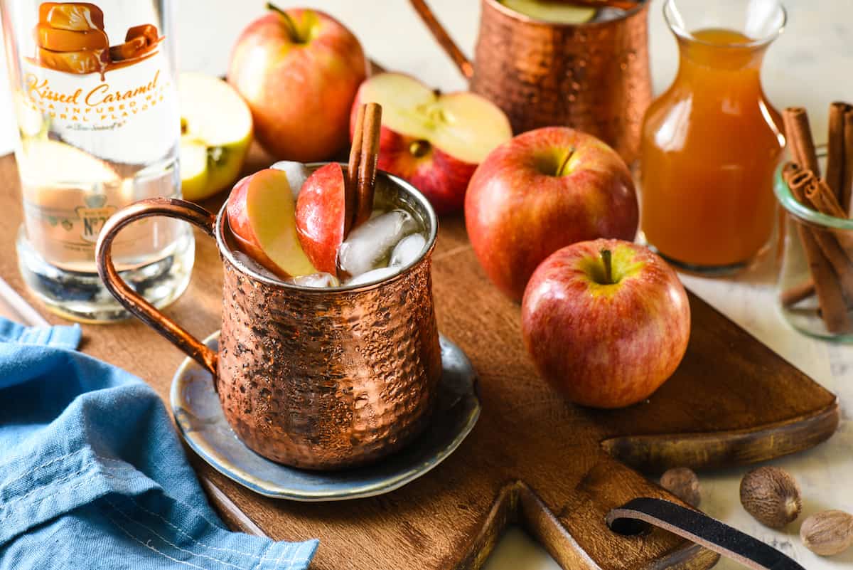 Wooden cutting board topped with apples and copper mugs filled with apple cider mule cocktails.