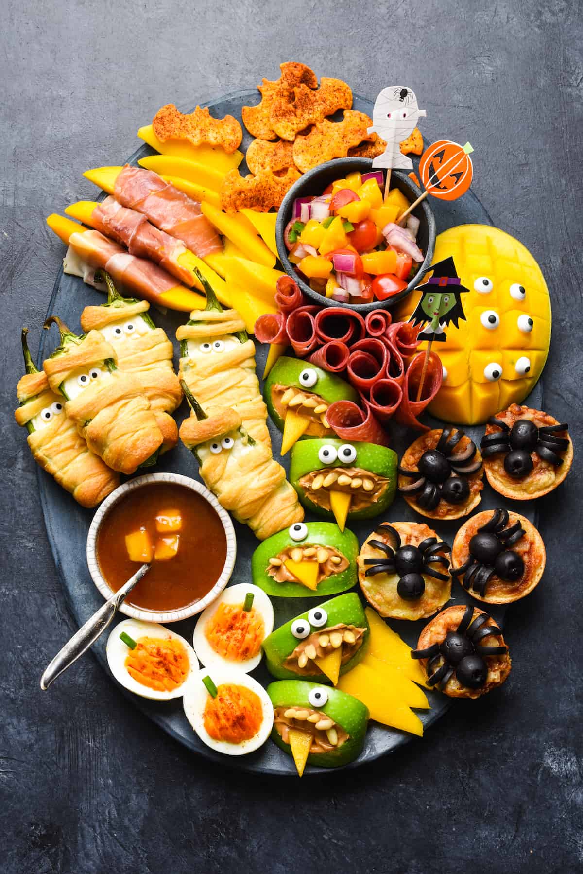 Spooky Halloween food ideas, like jalapeno popper mummies, apple monsters, bat tortilla chips and spider mini pizzas, arranged on a large dark platter on a dark background.