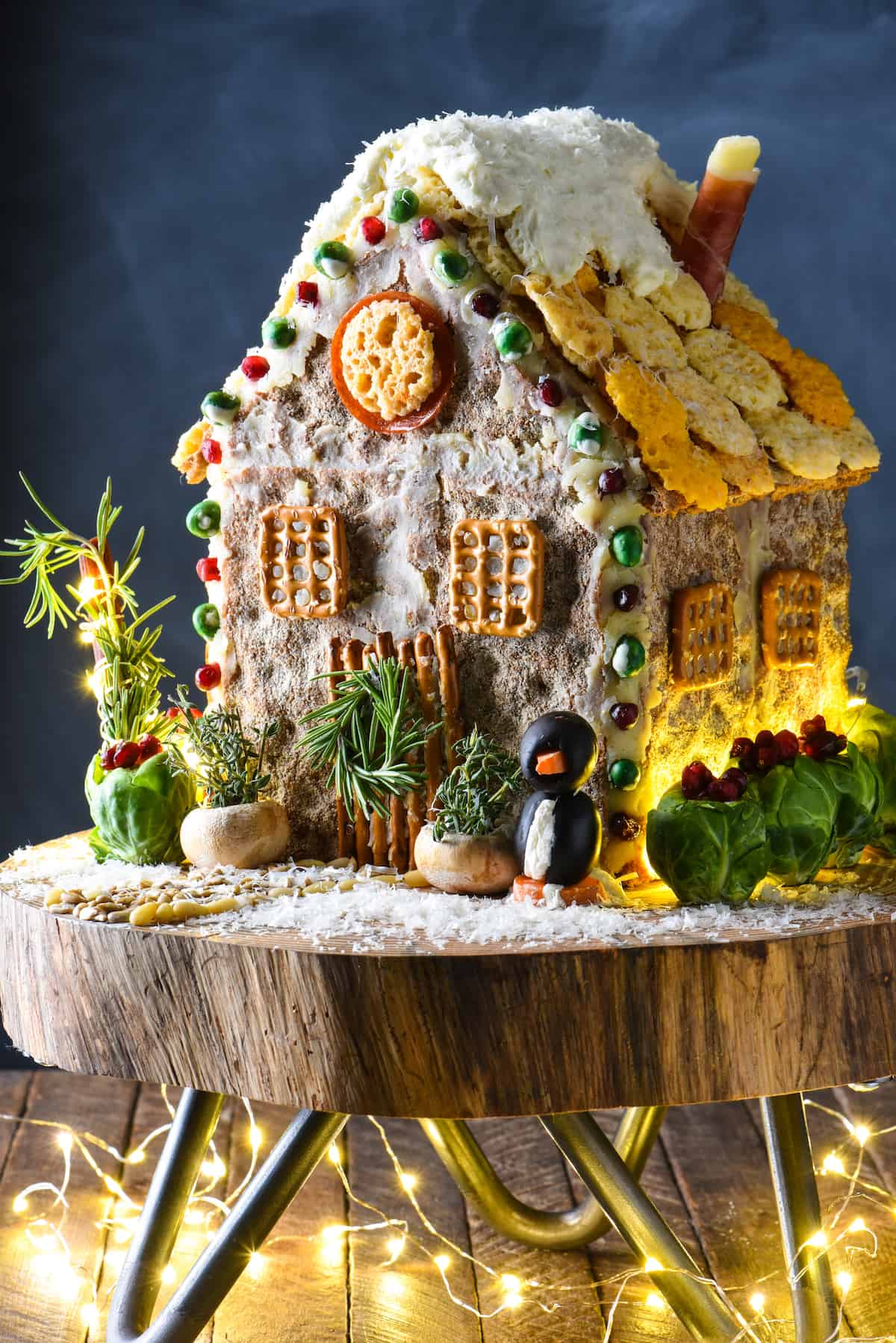 A savory gingerbread house (charcuterie chalet) on a wooden stand, with holiday lights decorating the scene.