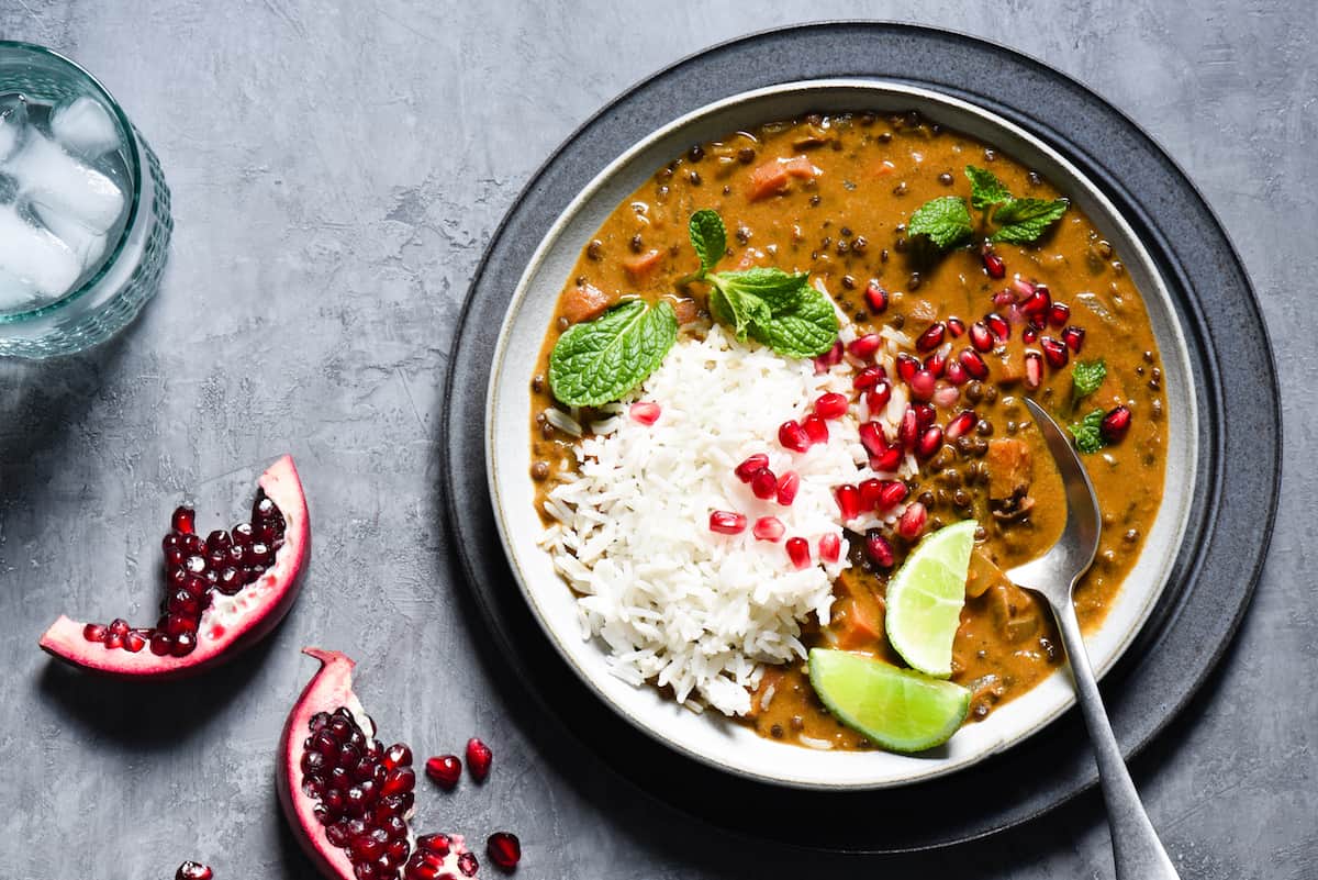Bowl of vegan curry and white rice garnished with lime wedges, pomegranate seeds and mint leaves.