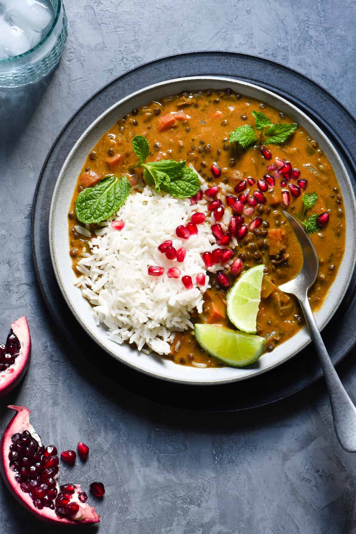 Bowl of vegan lentil curry and white rice garnished with lime wedges, pomegranate seeds and mint leaves.