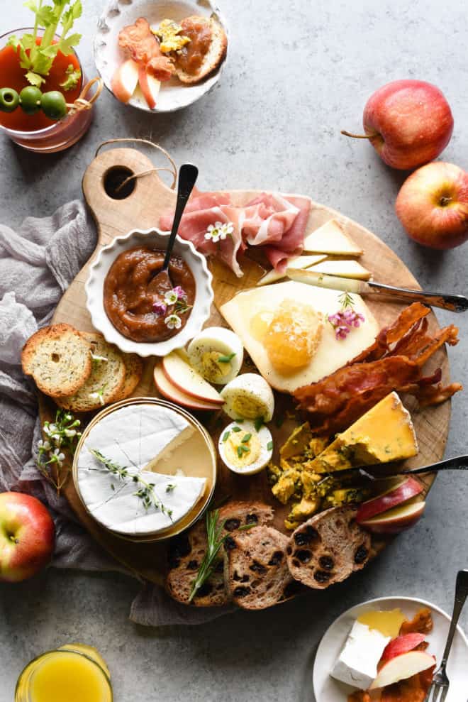 Round wooden cutting board filled with brunch menu ingredients like cheese, hard boiled eggs, bread, apple butter, bacon and prosciutto.