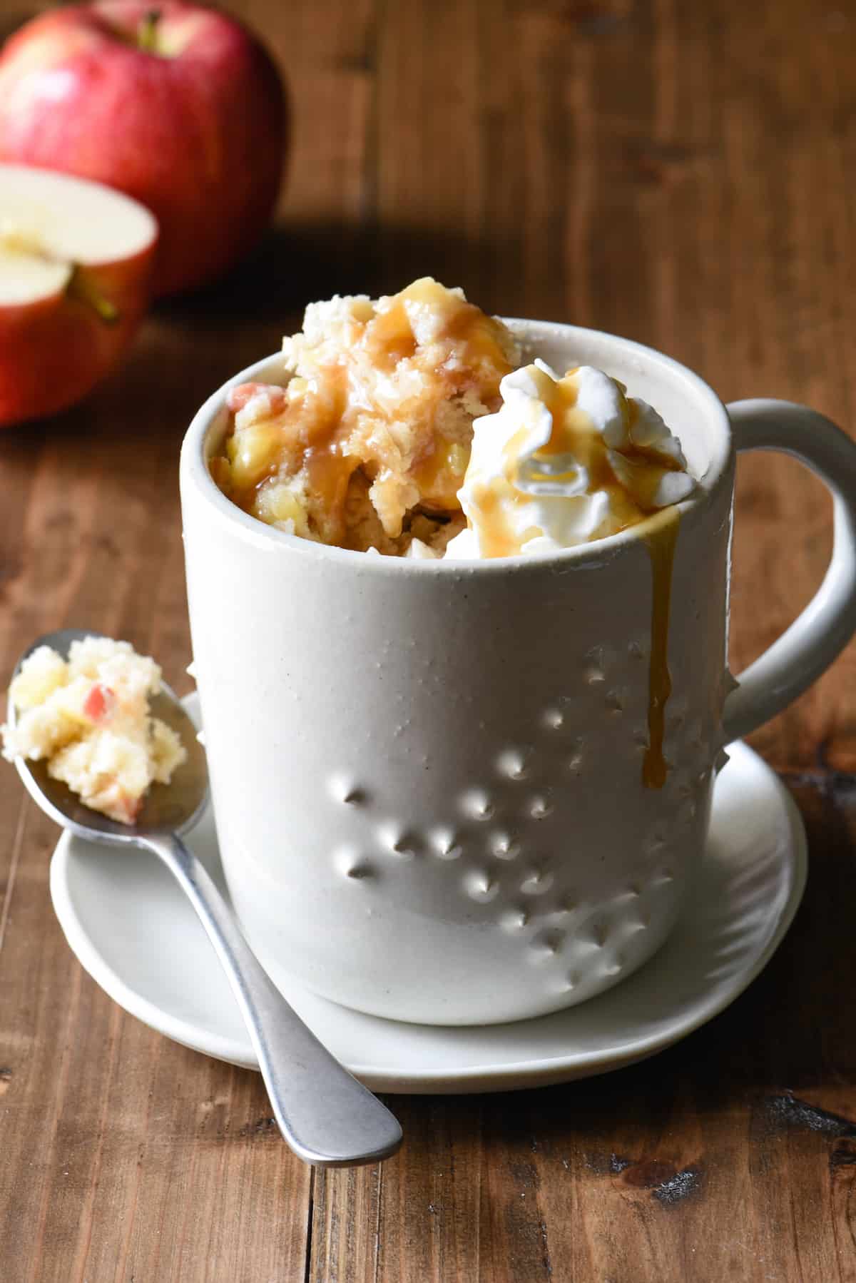 Textured ceramic mug filled with apple mug cake, topped with whipped cream and caramel.