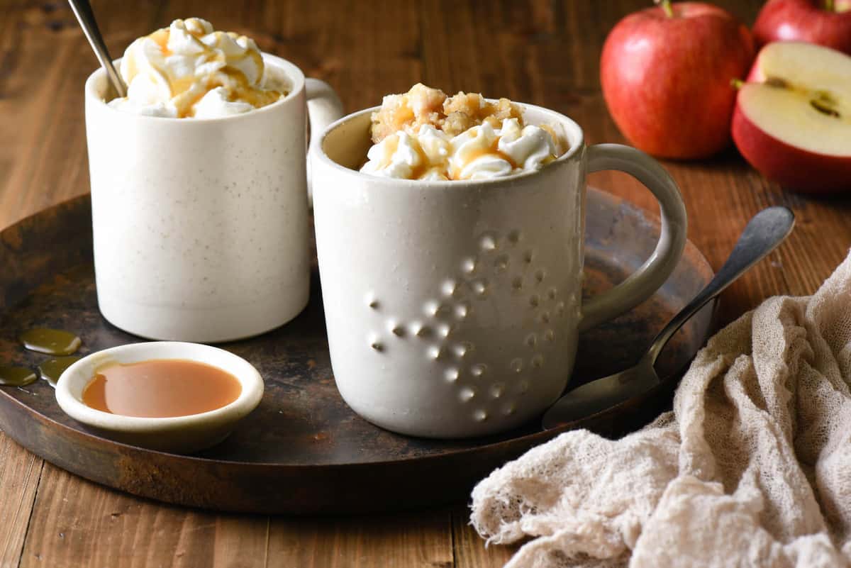 Small rustic round tray topped with two mugs filled with an easy microwave dessert cake and a small bowl of caramel sauce.