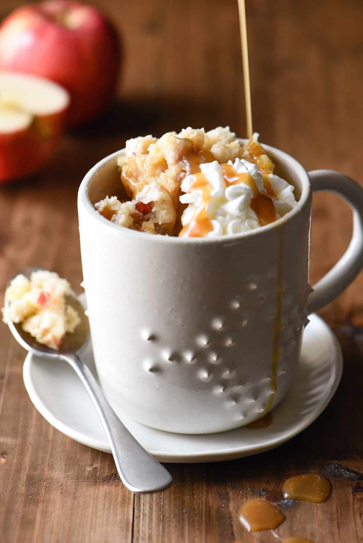 Textured ceramic mug filled with apple mug cake, topped with whipped cream. Caramel being drizzled over mug.