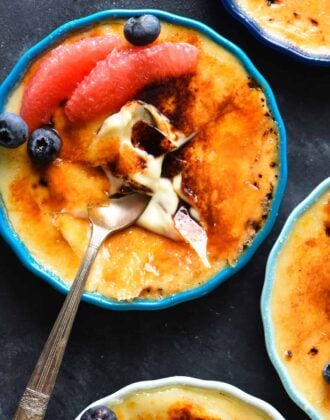Grapefruit creme brulee, garnished with grapefruit segments and blueberries, on a dark surface, with a spoon digging into it.