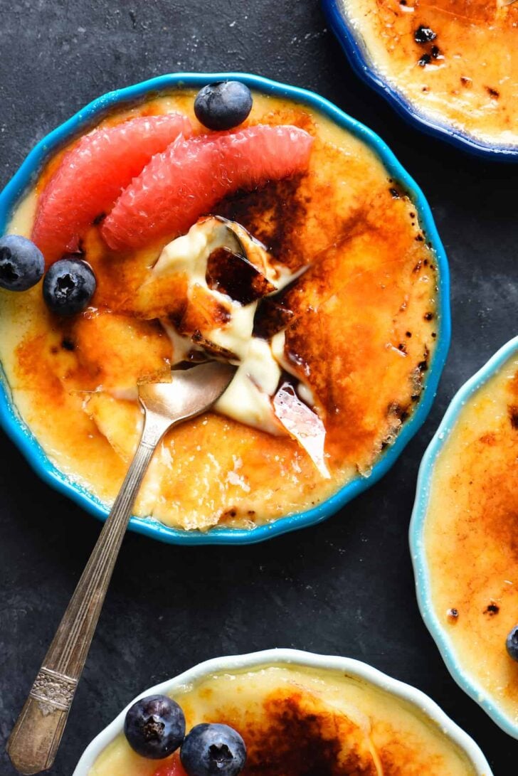 Grapefruit creme brulee, garnished with grapefruit segments and blueberries, on a dark surface, with a spoon digging into it.