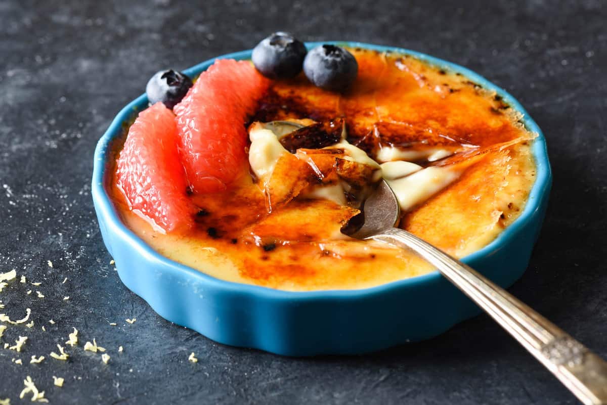 A blue ramekin of custard with crunchy torched sugar on top, with a spoon digging into it. Grapefruit segments and blueberries garnish the custard.