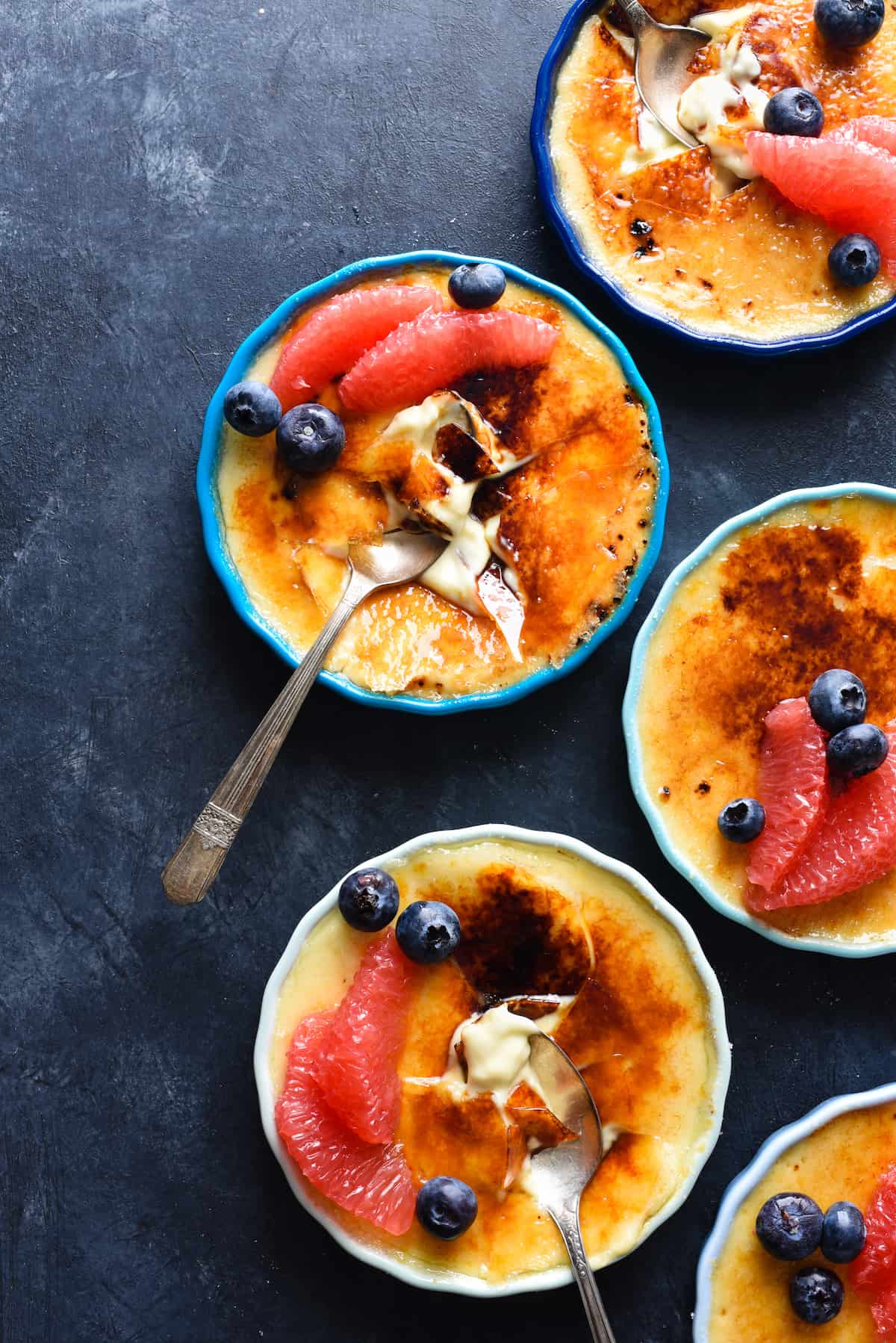 Grapefruit creme brulee in ramekins, garnished with grapefruit segments and blueberries, on a dark surface, with a spoons digging into them.