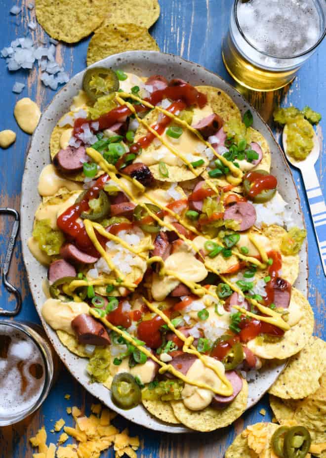 A large platter of nachos made with tortilla chips, cheese sauce, sliced hot dogs and pickled jalapenos.