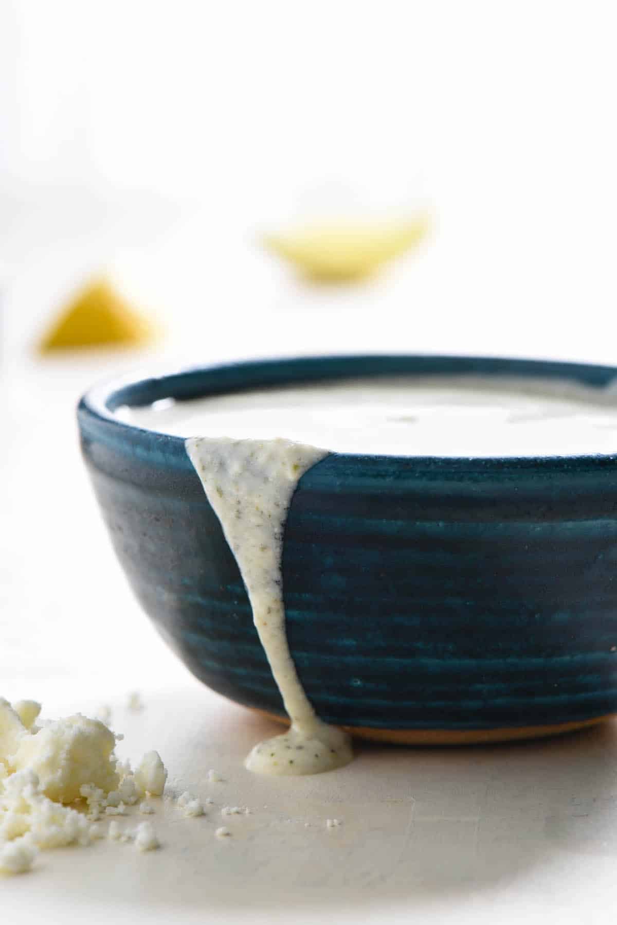 Blue pottery bowl filled with feta salad dressing, with some spilling over side. Crumbled feta next to bowl.