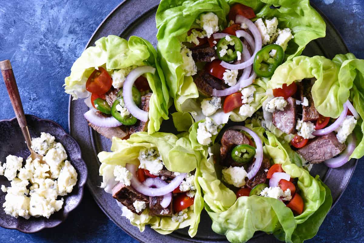 Steak lettuce wraps garnished with blue cheese and vegetables on a blue background.