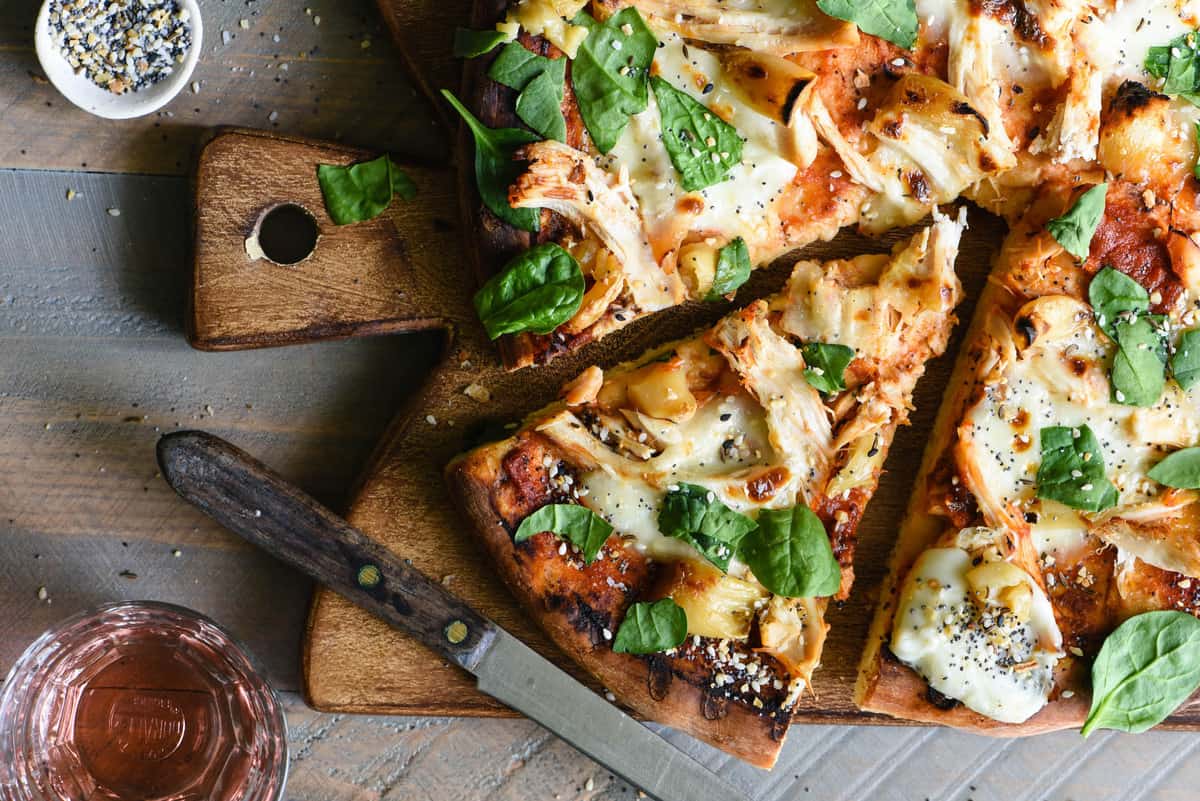 Overhead photo of pizza with chicken, roasted garlic and greens, on cutting board with knife.