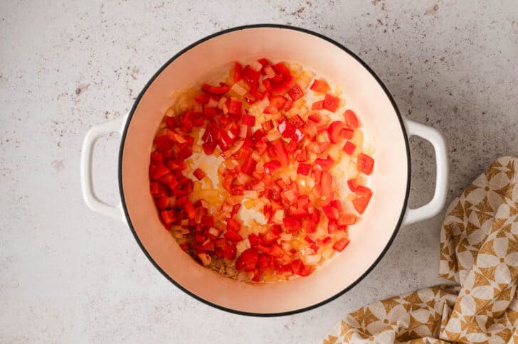 Red bell pepper and onion being cooked in a white Dutch oven.