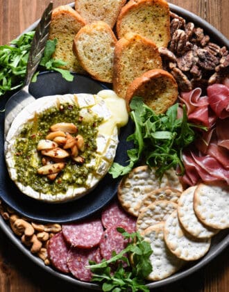 Savory baked brie topped with pesto and roasted garlic cloves, on a platter with crackers and charcuterie.