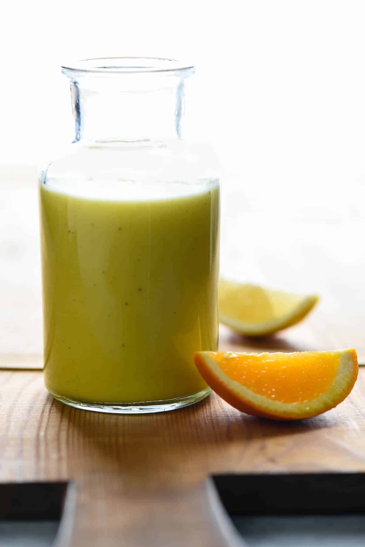 Small glass bottleneck jar filled with a yellow-hued healthy salad dressing. Slices of orange and lemon on cutting board near dressing.