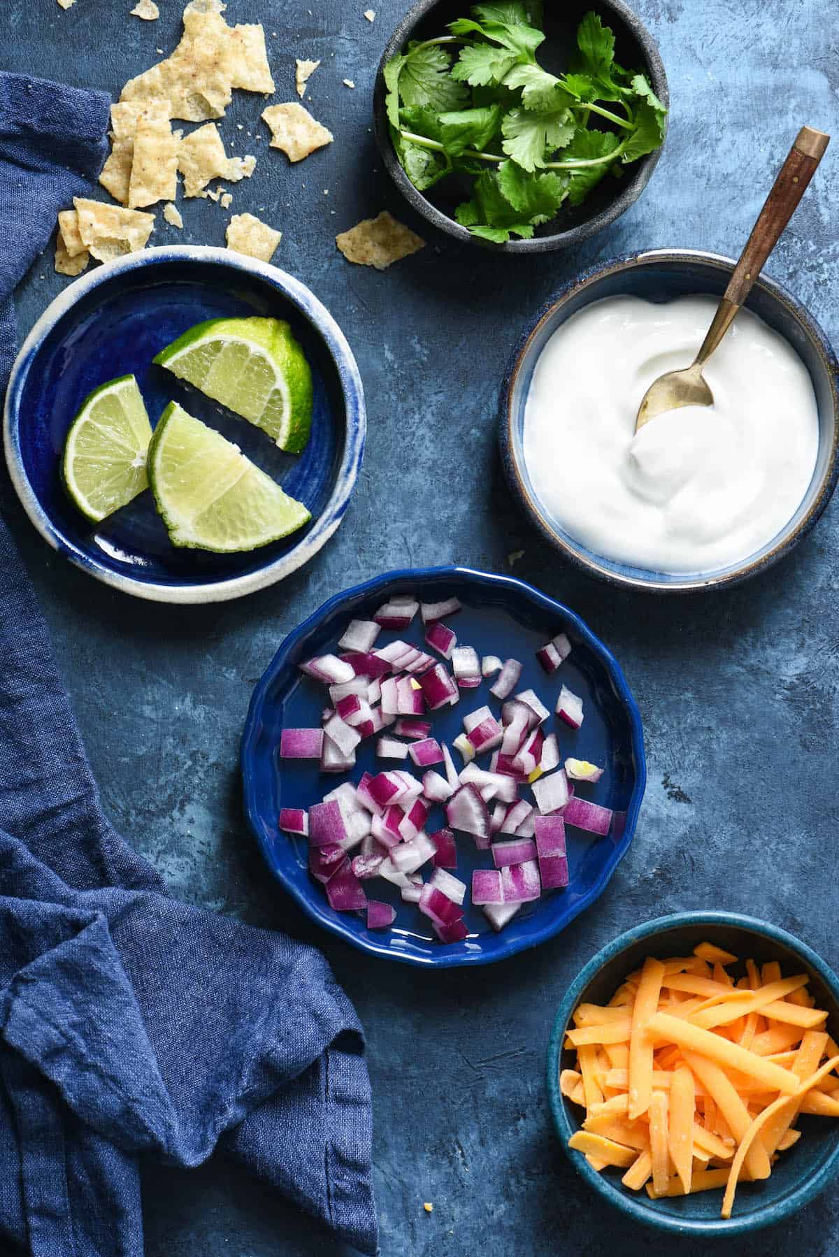 Bowls on blue background filled with garnished for taco soup, including cilantro, limes, sour cream, red onion and shredded cheese.