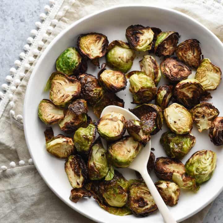 Air fried brussels sprouts in shallow white bowl with white serving spoon, on top of beige linen with pom pom edging.