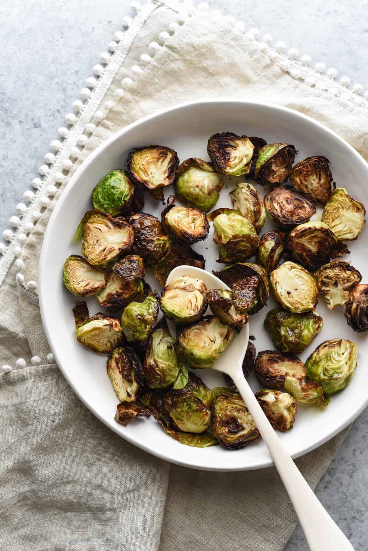Air fried brussels sprouts in shallow white bowl with white serving spoon, on top of beige linen with pom pom edging.