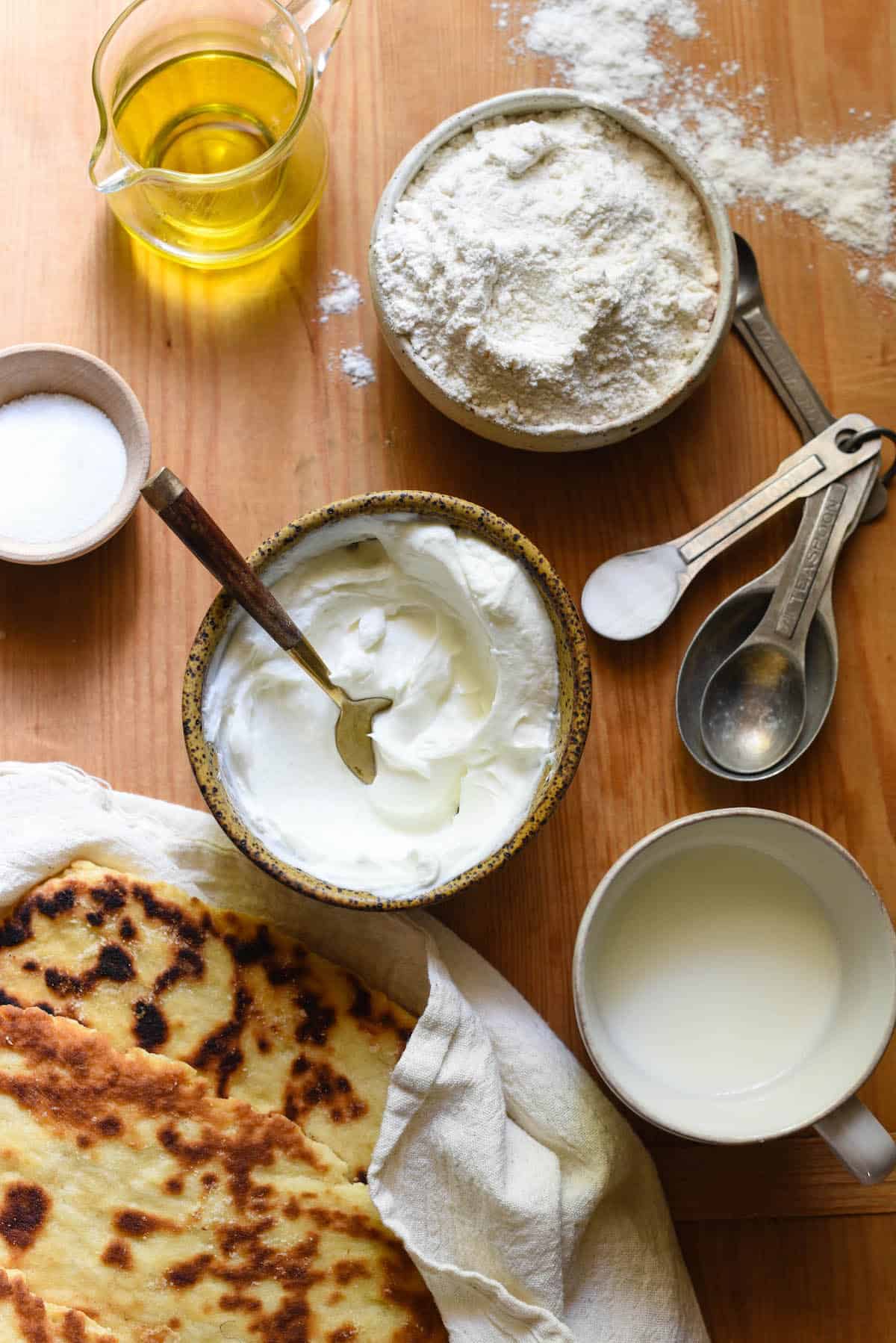 Overhead image of ingredients for homemade flat bread - olive oil, flour, baking soda, yogurt, milk and sugar. Finished flatbreads alongside.