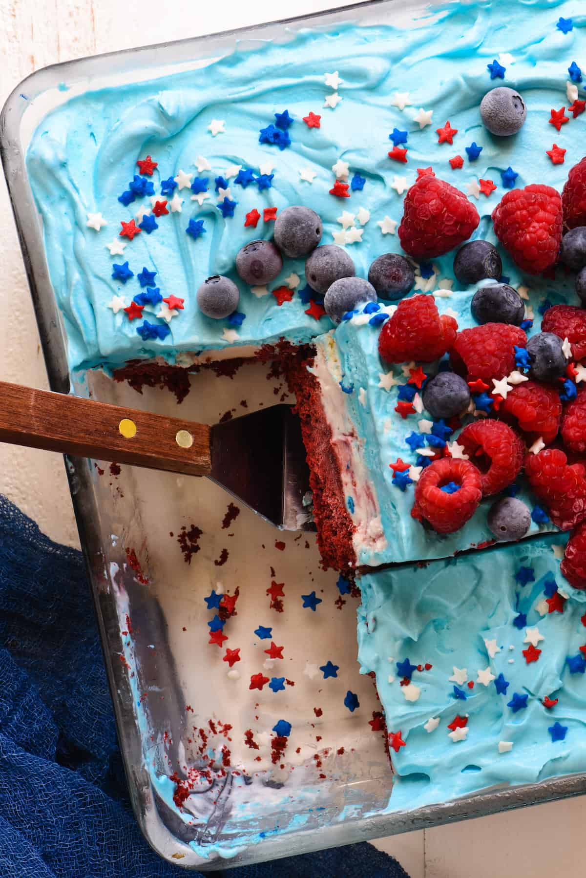 Closeup of pan of red white and blue frozen cake topped with berries. One piece cut and tilted, resting on spatula.