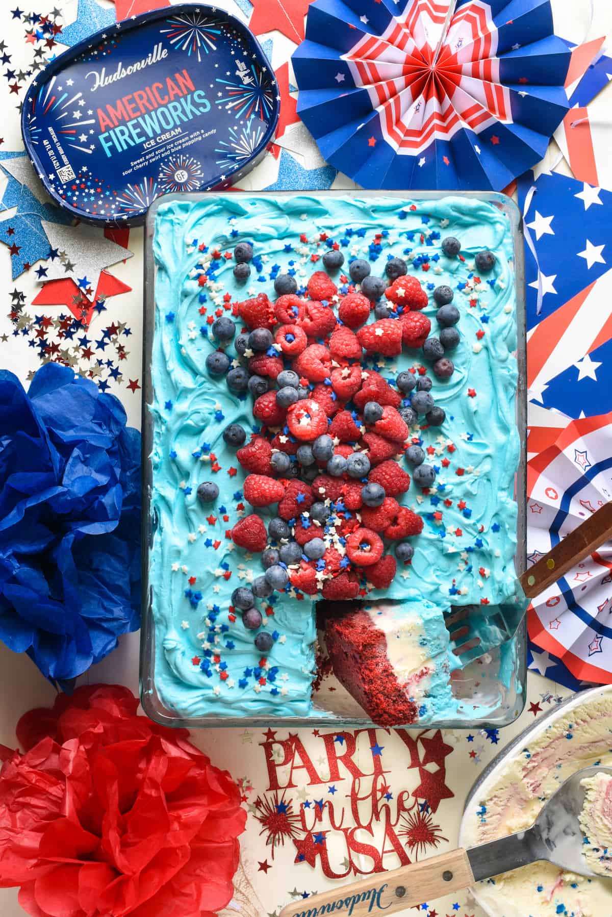 Overhead shot of red white and blue ice cream cake with one piece cut and turned. Cake is topped with berries and sprinkles.