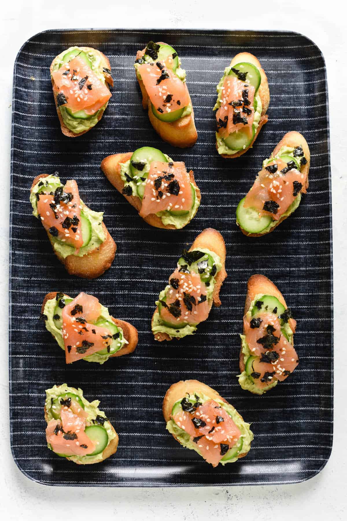 Avocado, salmon and cucumber crostini arranged on blue and white striped tray.