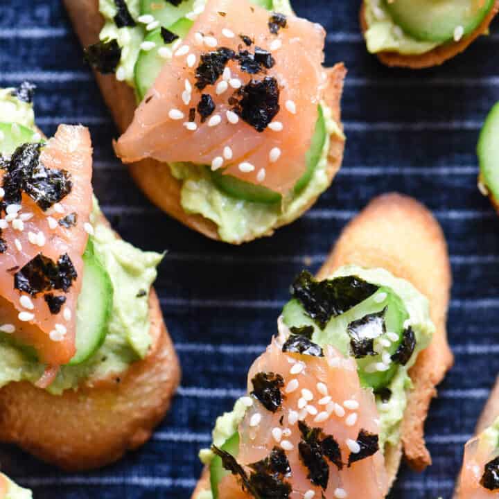 Toasted baguette topped with avocado spread, cucumber and smoked salmon, on top of blue and white striped serving tray.
