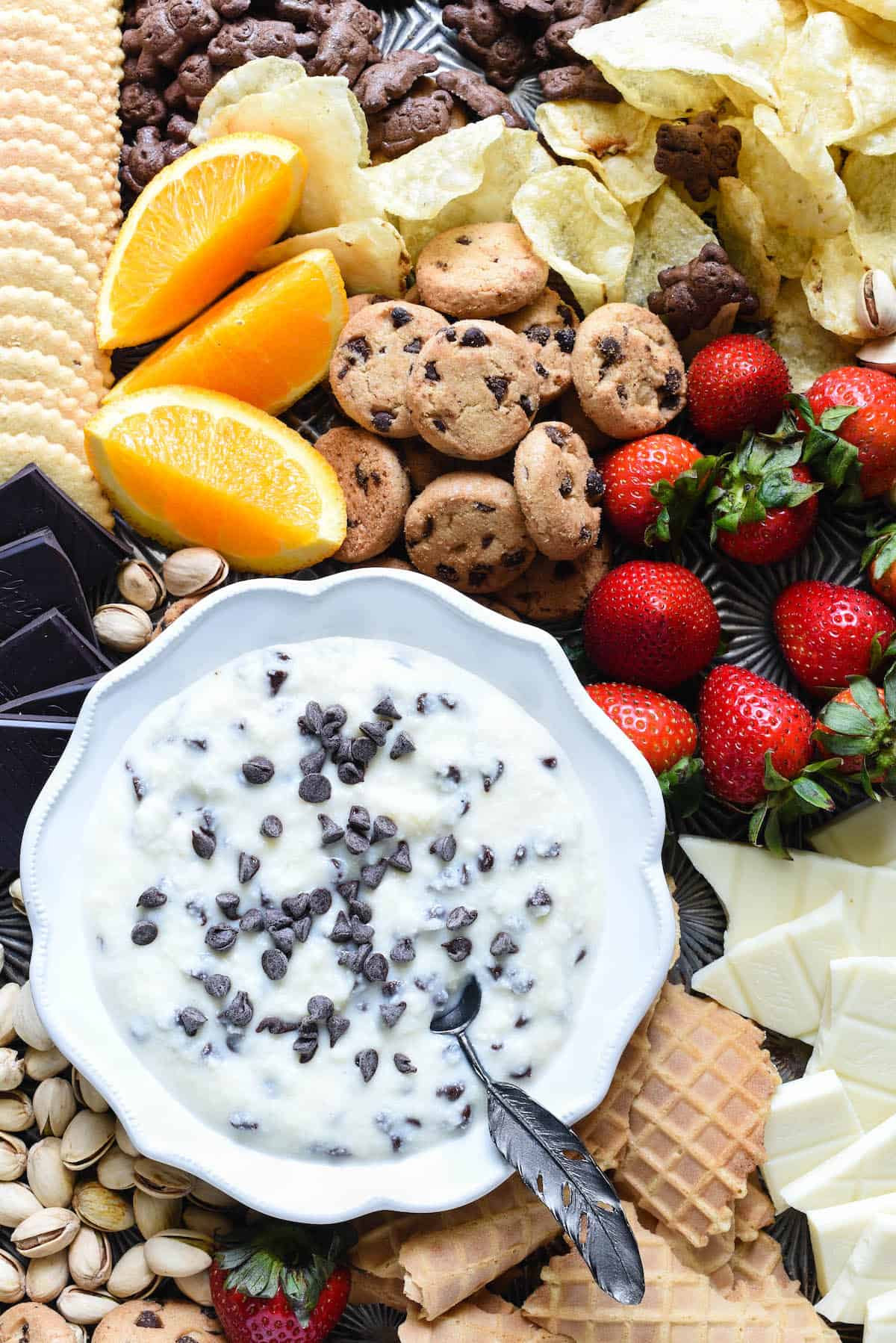 Ricotta and mascarpone mixture topped with mini chocolate chips in fluted white bowl, surrounded by cookies, fruit, chocolate and nuts.