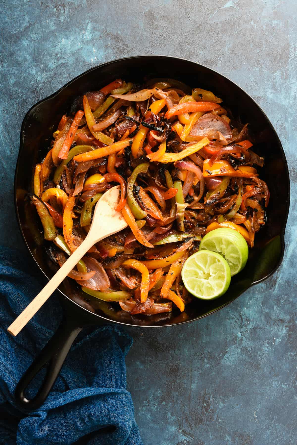 Cast iron skillet filled with cooked fajita veggies, with wooden spoon and lime halves.