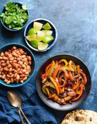 Bowls of sauteed fajita veggies, pinto beans, lime wedges and cilantro on blue background with charred tortillas.