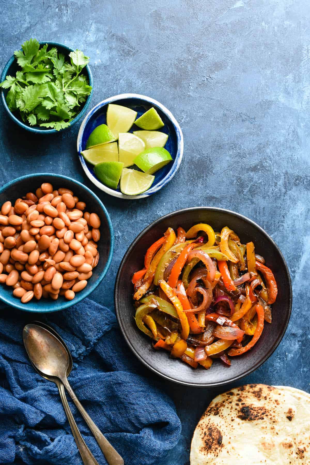 Bowls of sauteed fajita veggies, pinto beans, lime wedges and cilantro on blue background with charred tortillas.