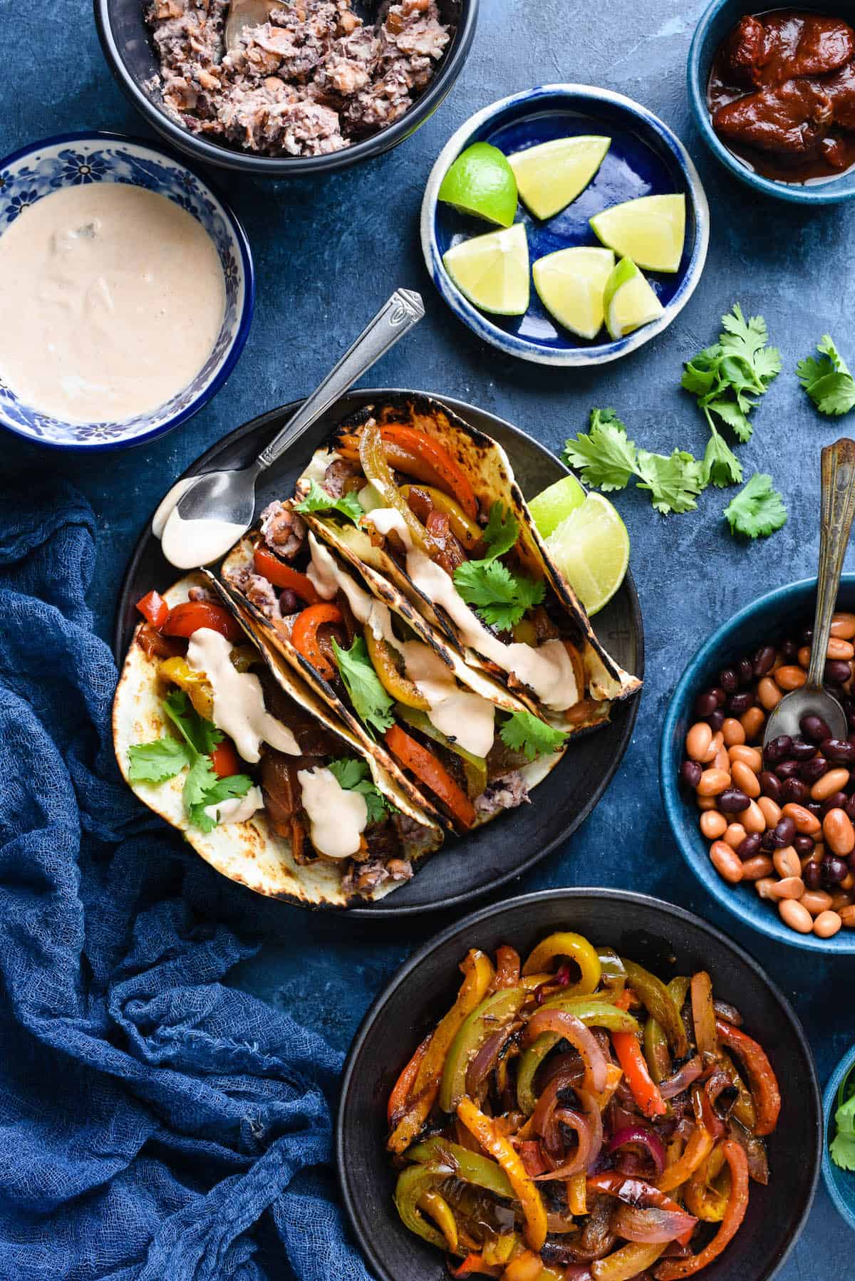 Overhead photo of plate of vegetarian fajijtas, along with bowls of creamy chipotle sauce, mashed beans, lime wedges, chipotles in adobo sauce, whole black beans and pinto beans, and cooked fajita vegetables.