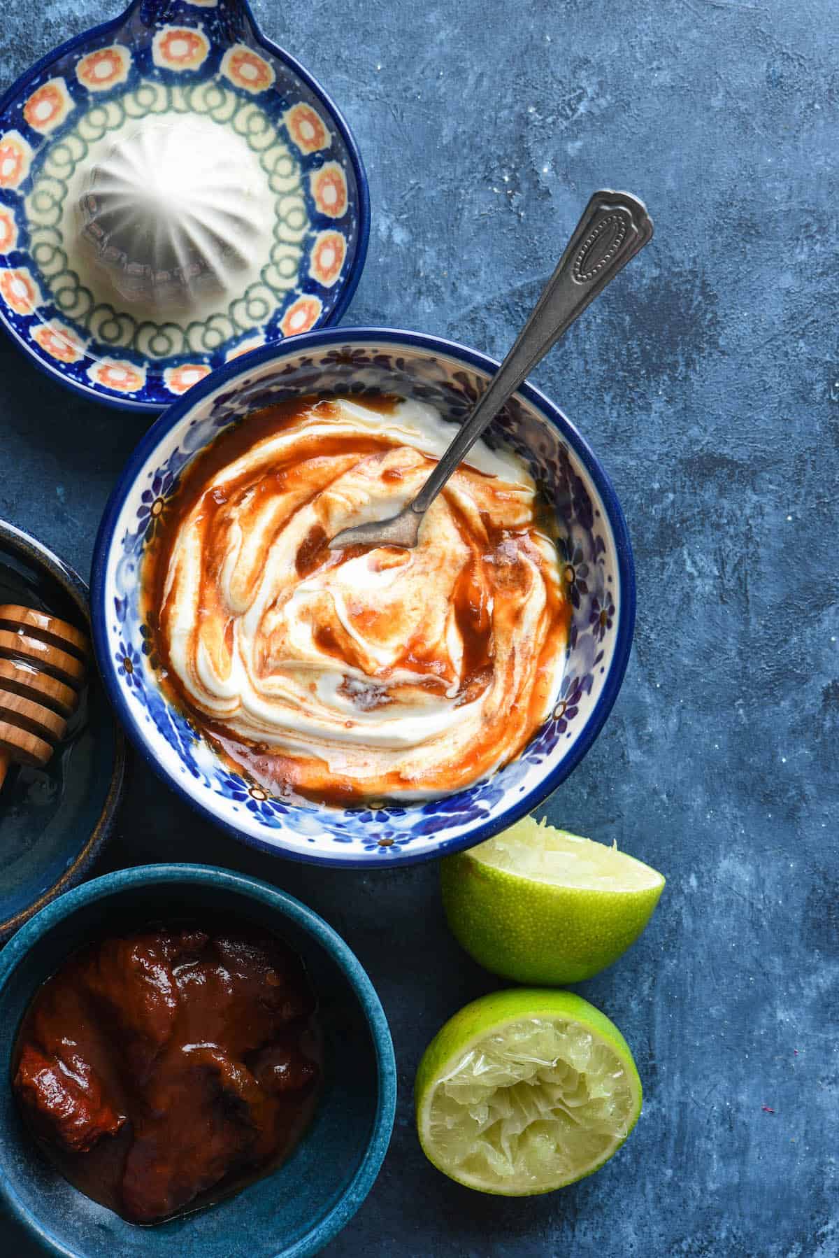 Bowl of creamy chipotle sauce on blue background with chipotles and limes alongside.