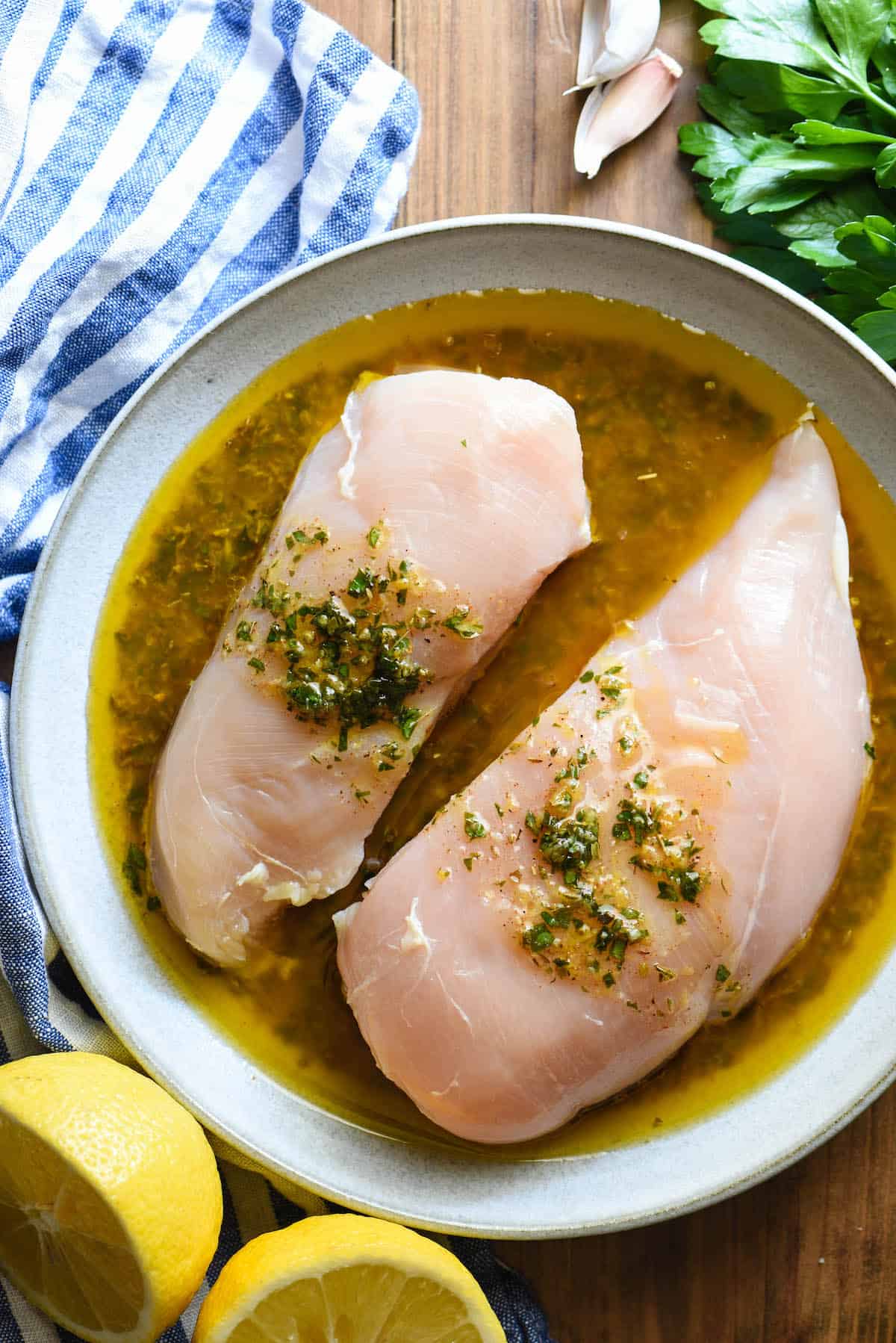 Raw chicken breasts in bowl of Greek chicken marinade. Blue and white napkin, cut lemon, herbs and garlic cloves on table near bowl.