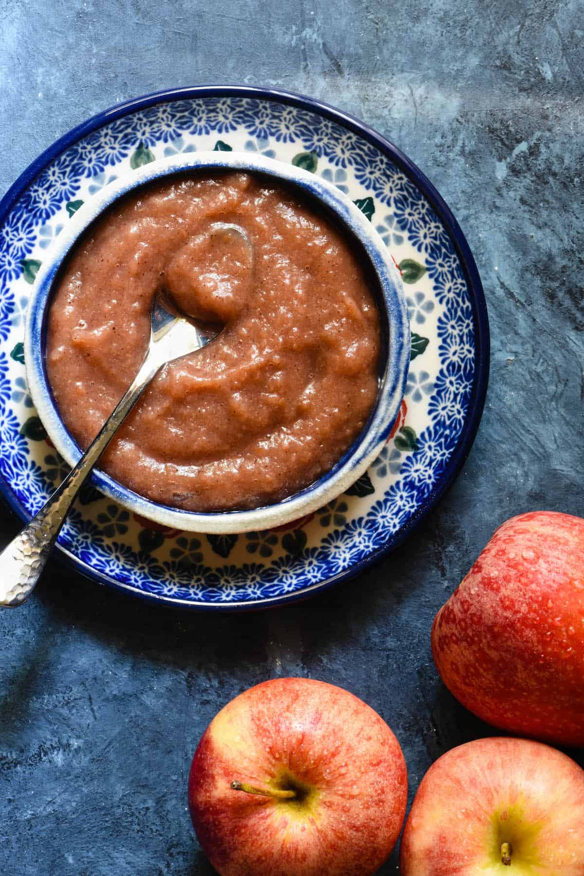 Pureed apple mixture in small dish on blue background with spoon. Fresh apples nearby on blue table.