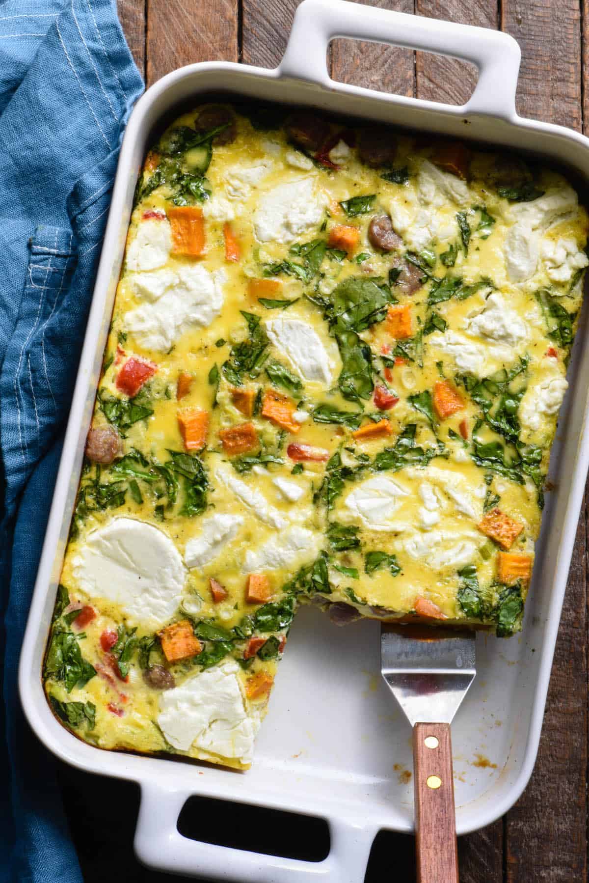 Sweet potato breakfast casserole made with eggs, sausage, spinach and goat cheese, in rectangular white baking dish, with a serving spatula lifting a piece out.
