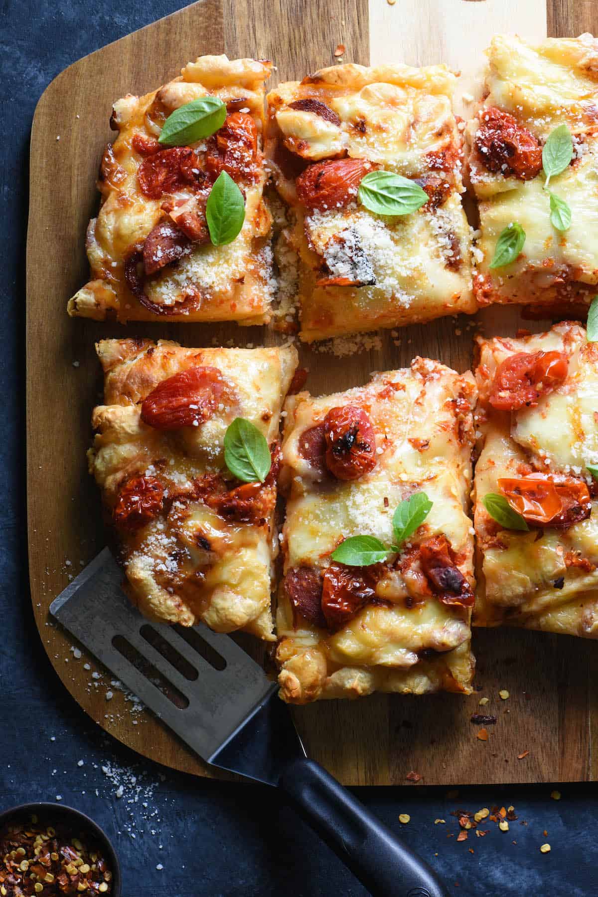 Closeup shot of Detroit style pizza topped with roasted tomatoes, parmesan cheese and basil, on wooden cutting board.