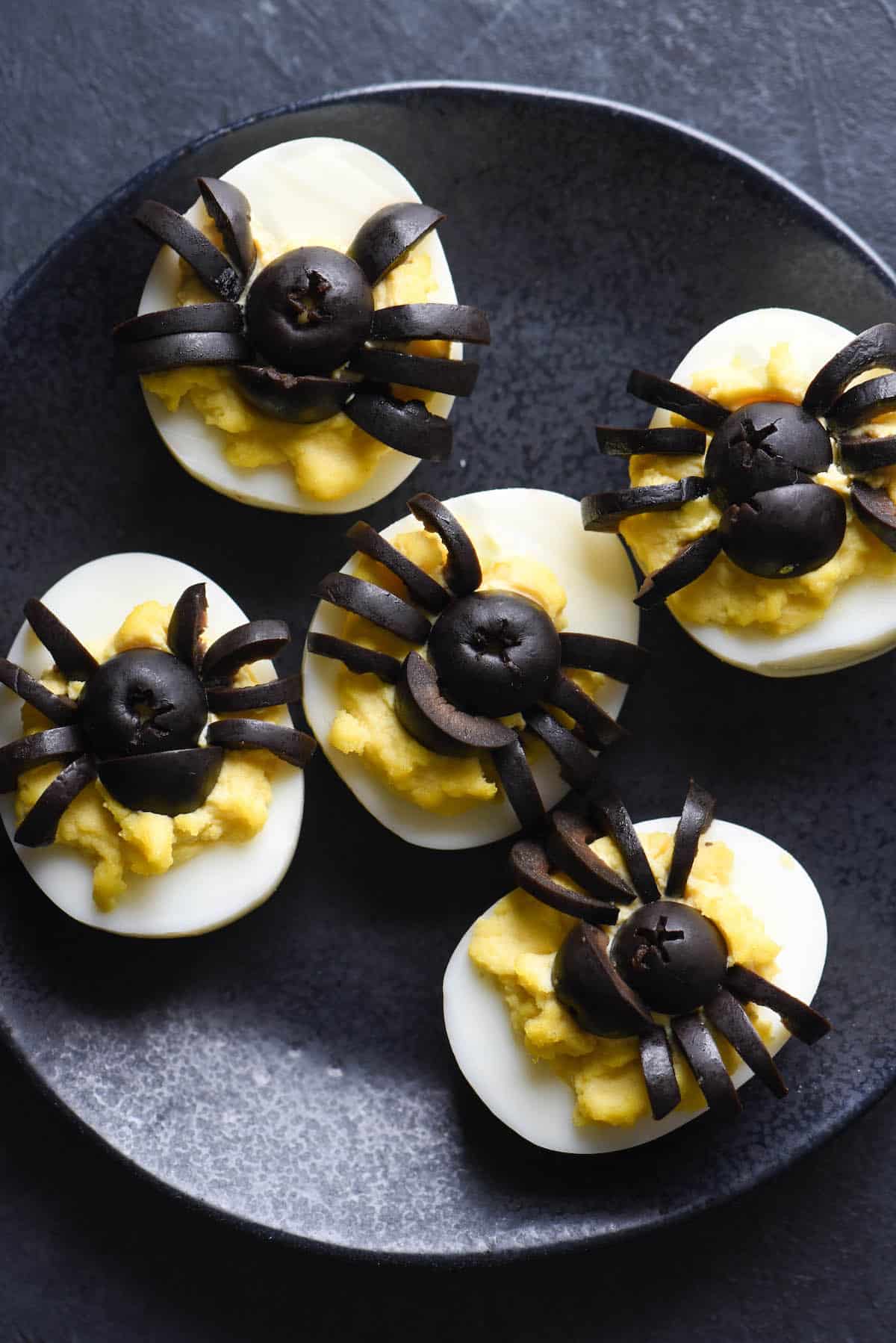 Black plate with deviled eggs on top. Black olives cut into the shape of spiders decorate the tops of the eggs.